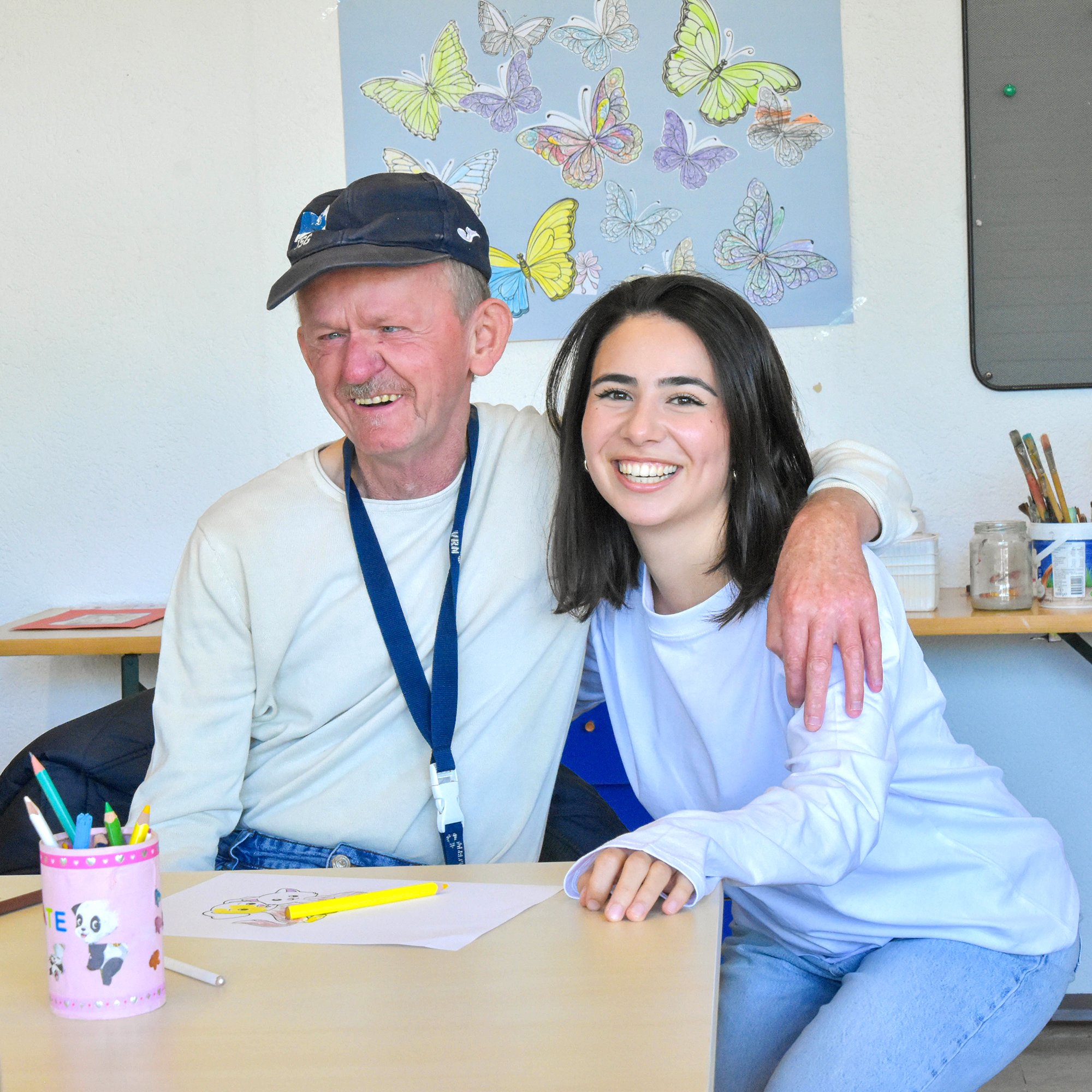 Ein älterer Mann und eine junge Frau sitzen nebeneinander an einem Tisch, sie lacht.