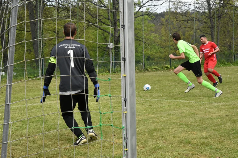 Männer spielen Fußball. Im Vordergrund ist ein Torwart mit dem Rücken zu sehen, zwei Spieler laufen mit Ball Richtung Tor.