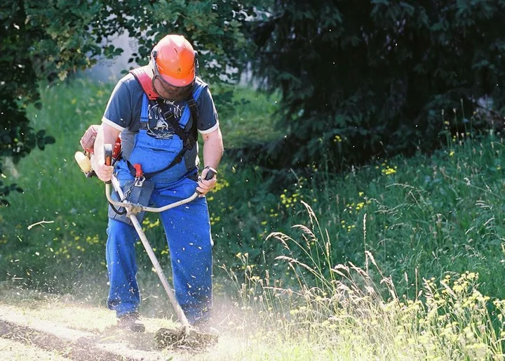 Ein Mann mit Arbeitskleidung mäht eine Wiese mit einem Freischneider.