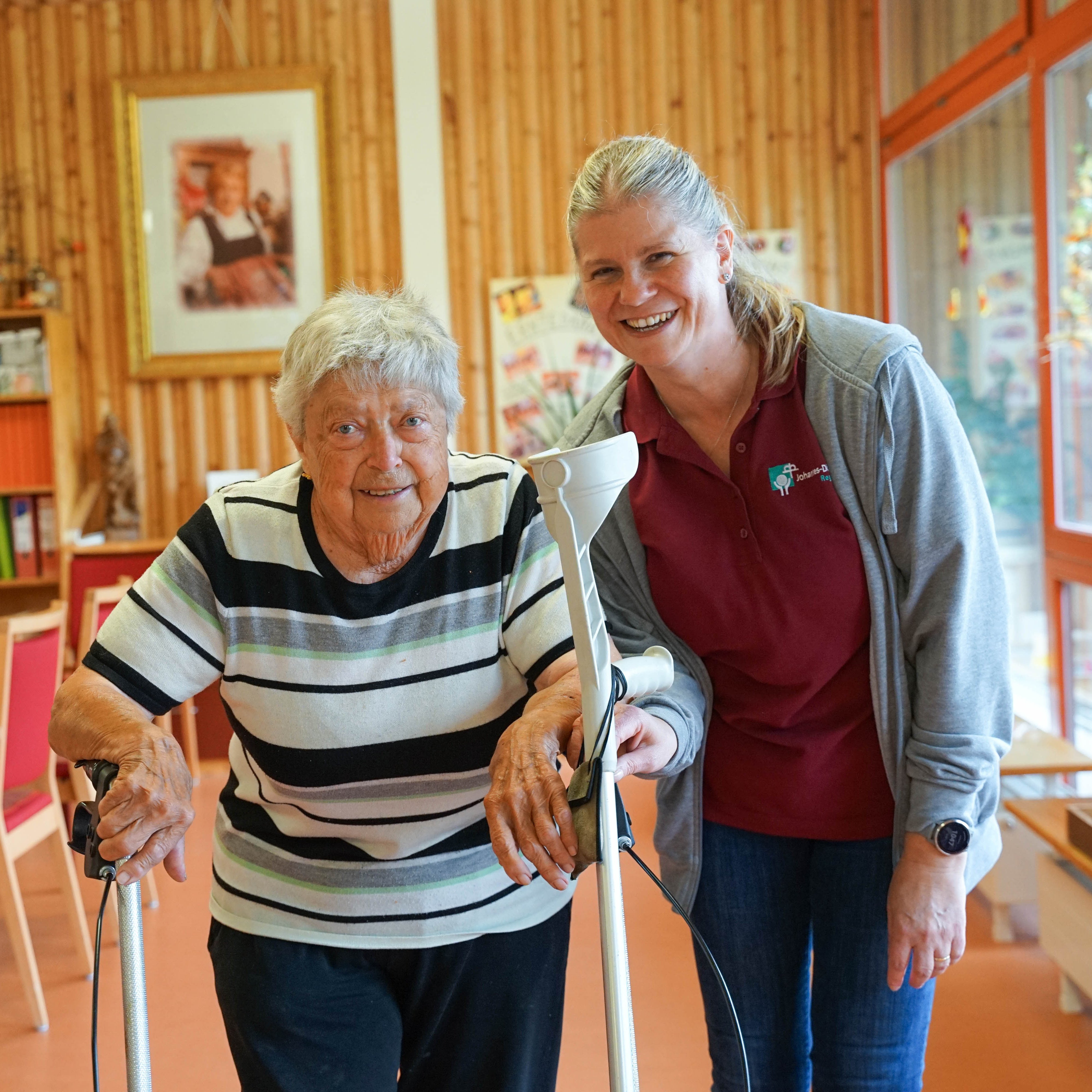 Eine ältere Frau läuft mit ihrem Rollator und eine Mitarbeiterin unterstützt sie.