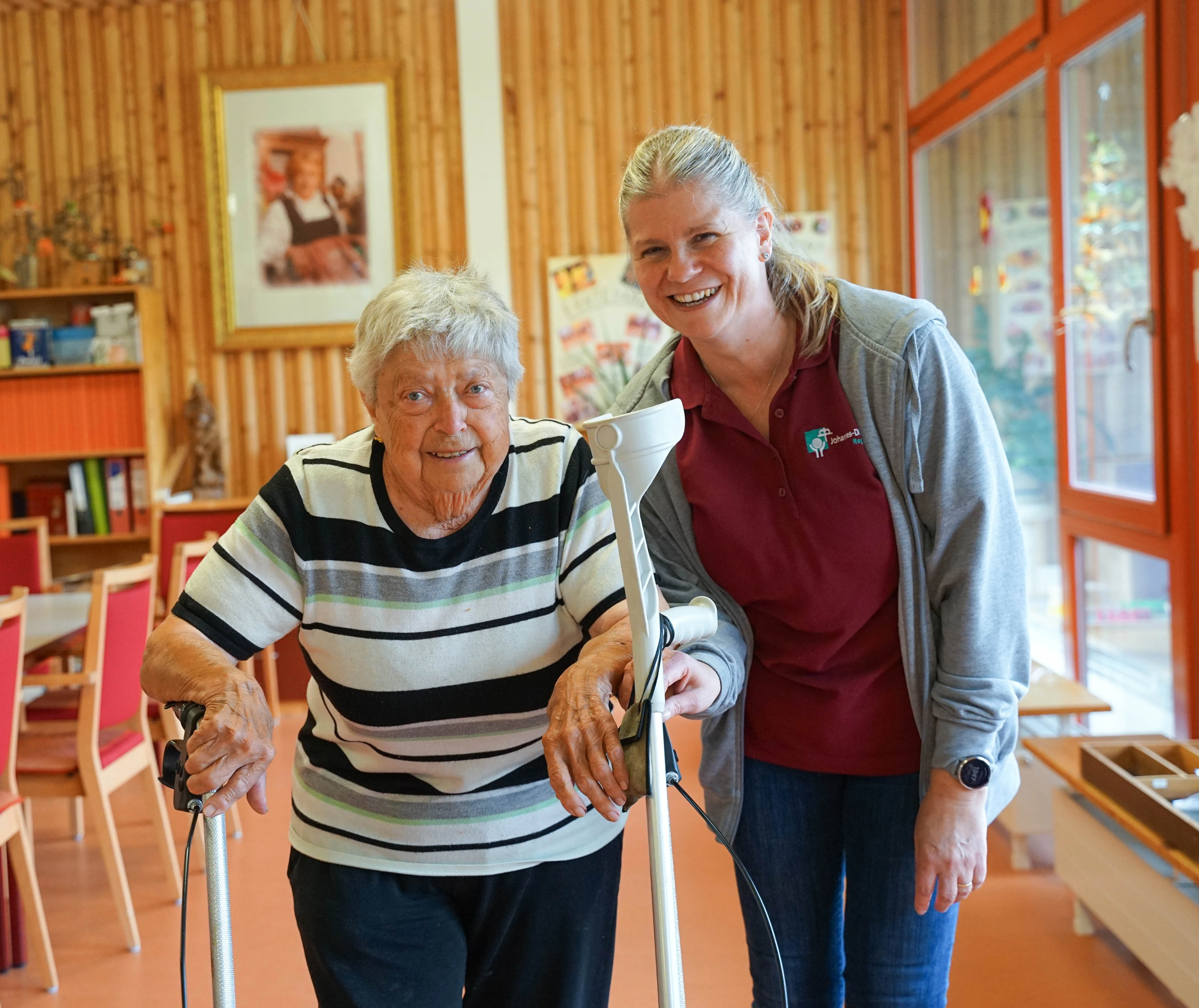 Eine ältere Frau läuft mit ihrem Rollator und eine Mitarbeiterin unterstützt sie.