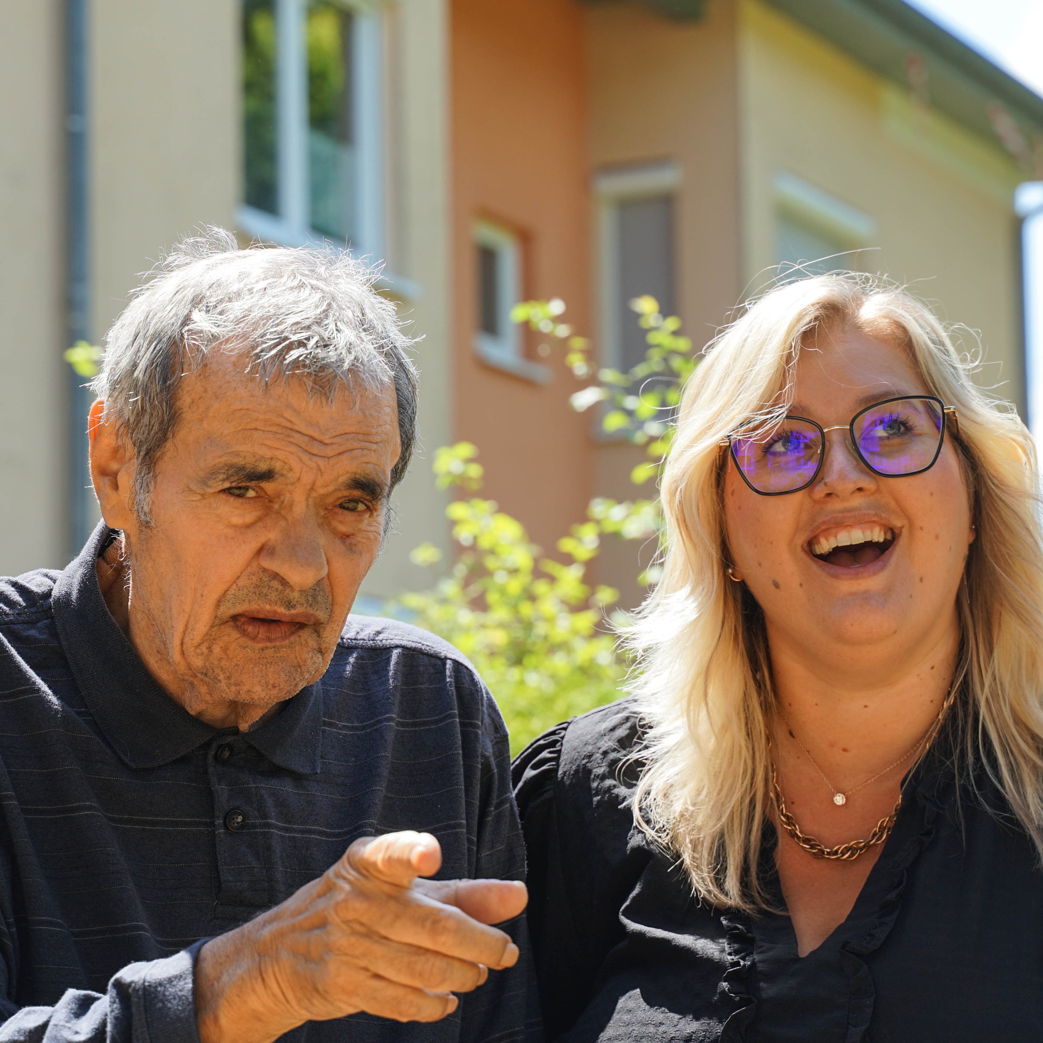 Ein älterer Mann zeigt mit dem Finger in eine Richtung, eine Frau sitzt neben ihm und lacht. Die Sonne scheint.