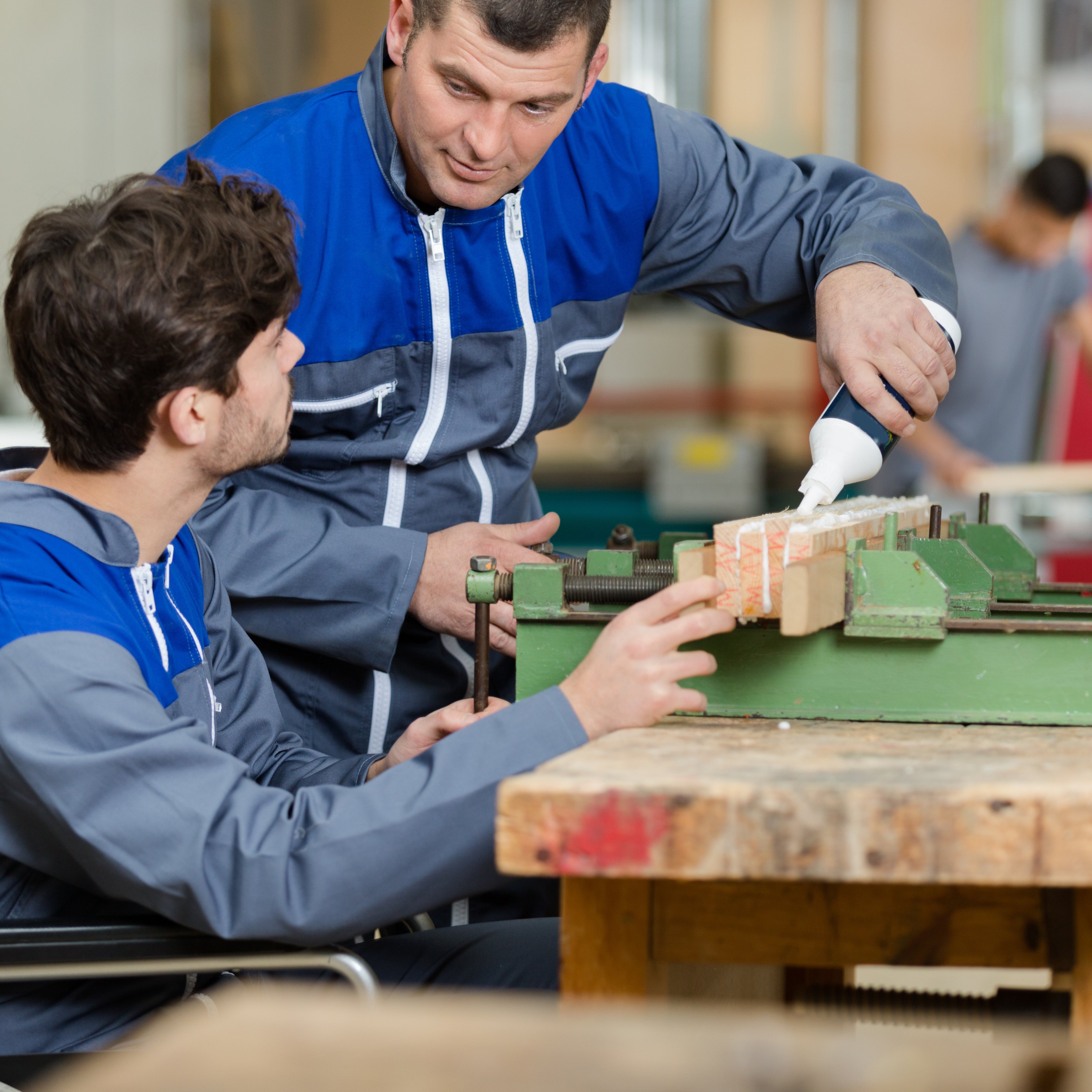 Ein junger Mann in einem Rollstuhl arbeitet mit Holz an einer Werkbank, ein Mann steht neben ihm und hilft dabei Bretter zu verleimen.