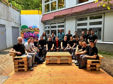 Audi-Auszubildende und Lehrkräfte der Johannesberg Schule sitzen auf einer Holzplattform vor dem Schulgebäude.