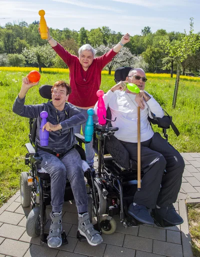 Zwei Menschen im Rollstuhl und eine Frau steht hinter ihnen. Alle halten einen farbigen Kegel in der Hand und lachen und jubeln, es ist Sommer.