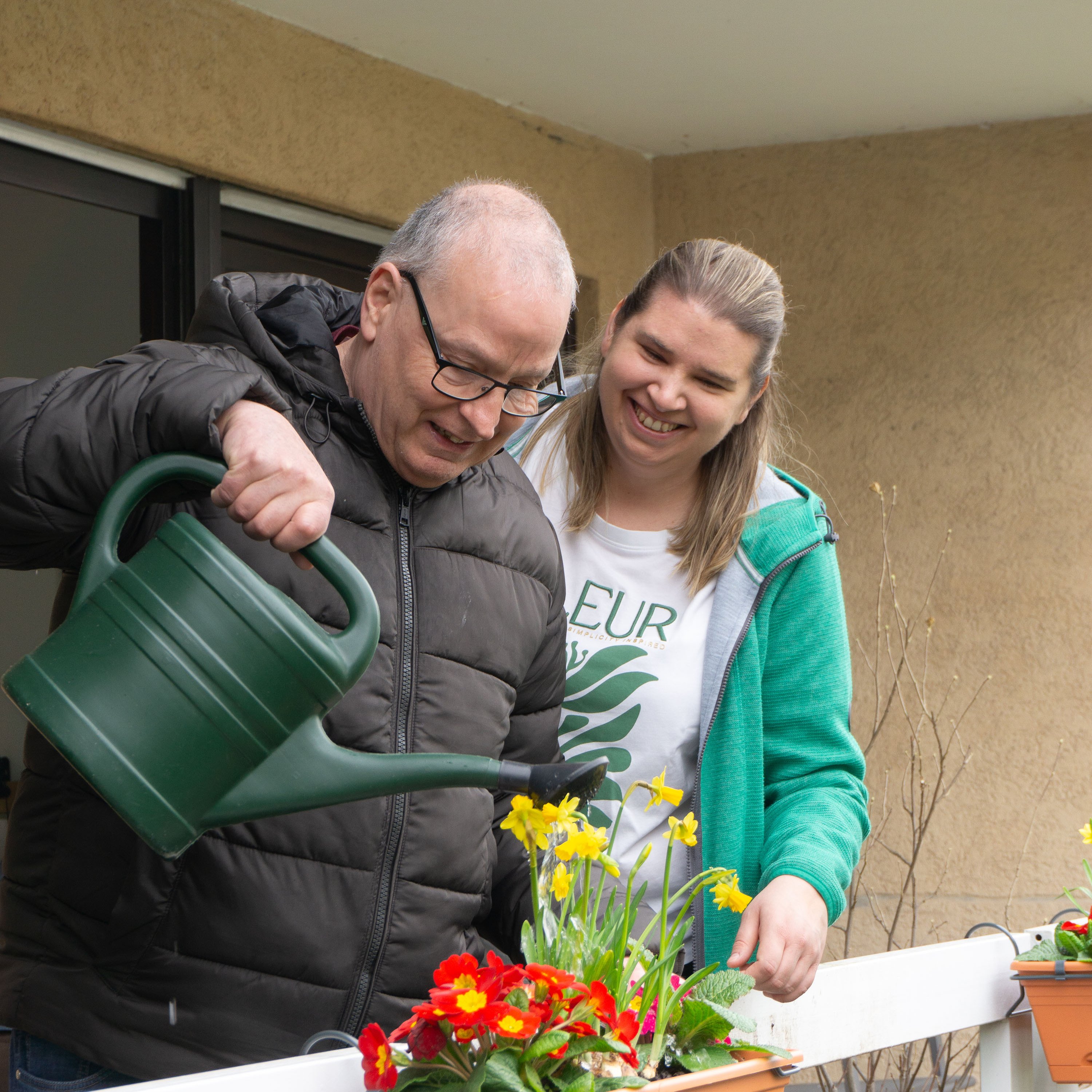 Eine Frau und ein Mann stehen auf einem Balkon. Vor ihnen befindet sich ein Blumenkasten mit Frühlingsblumen. Der Mann gießt diese mit einer dunkelgrünen Gießkanne.