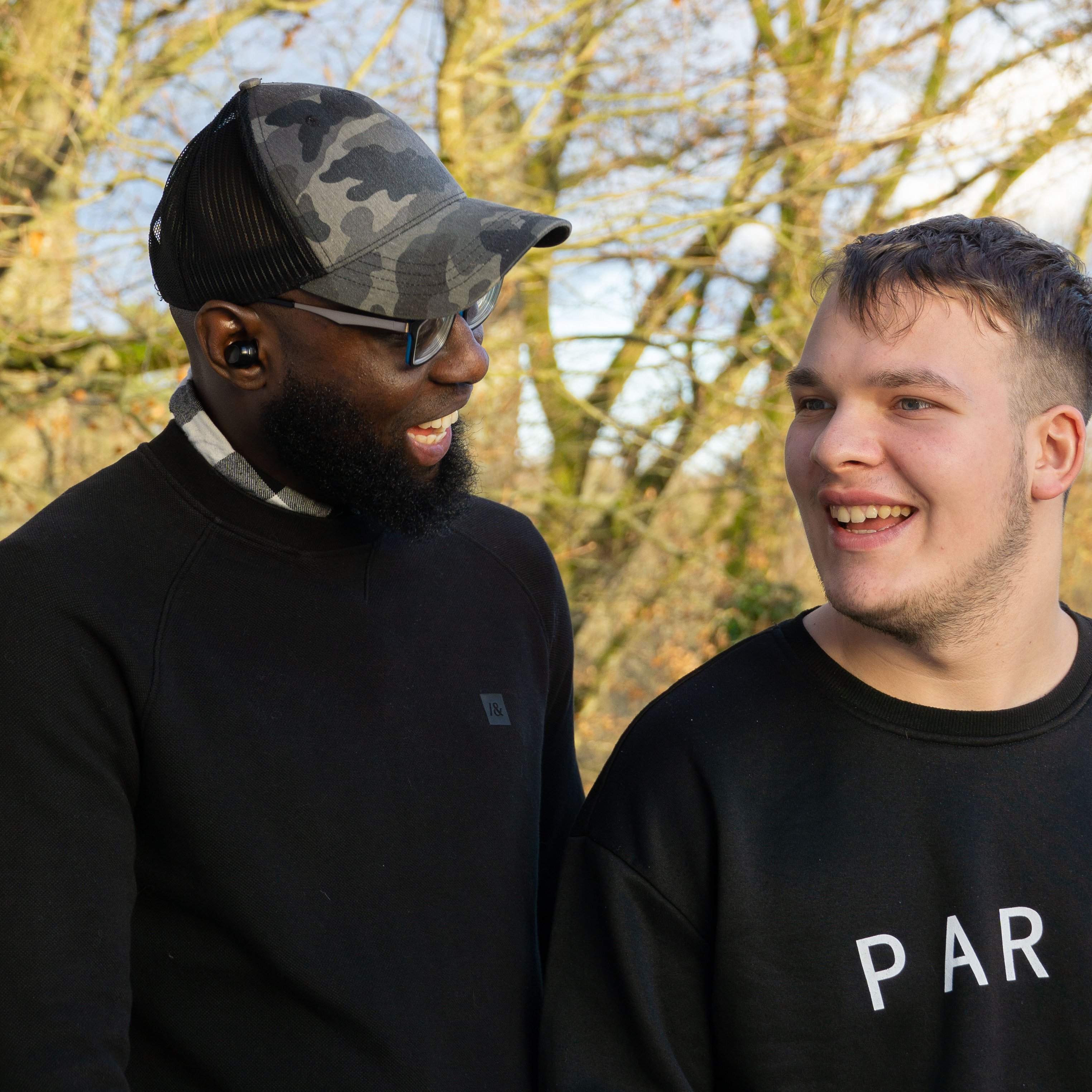 Ein Mitarbeiter und ein junger Klient lachen zusammen. Sie sind draußen, im Hintergrund sind Bäume zu sehen.