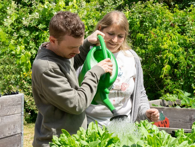 Eine Mitarbeiterin und ein Klient stehen im Garten an einem Hochbeet mit Pflanzen. Der Klient gießt die Pflanzen mit einer Gießkanne.