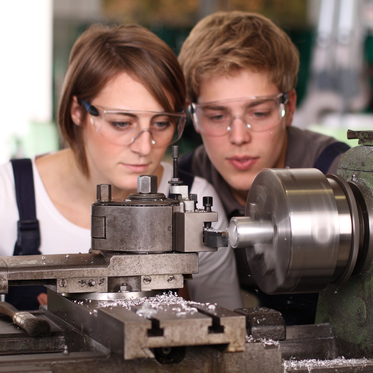 Eine junge Frau und ein junger Mann mit Schutzbrillen schauen auf eine Maschine, die im Vordergrund läuft und Metallsplitter verursacht. 