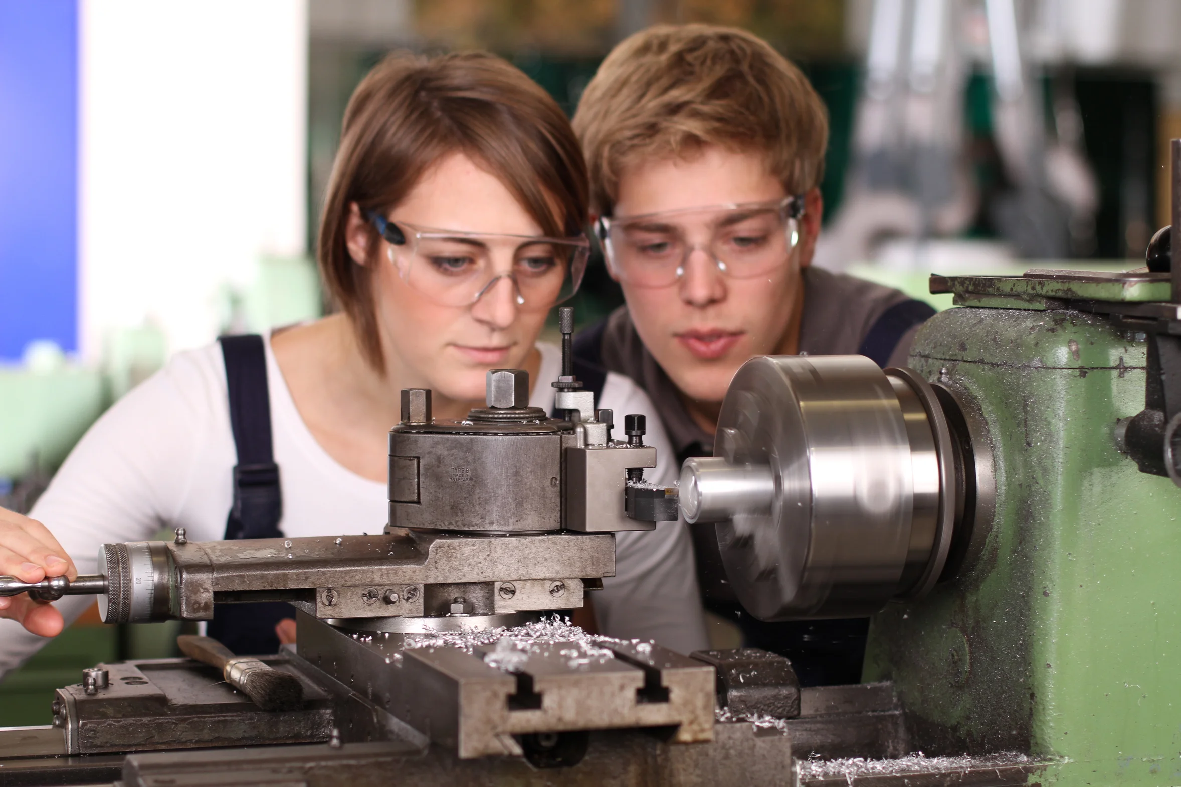 Eine junge Frau und ein junger Mann mit Schutzbrillen schauen auf eine Maschine, die im Vordergrund läuft und Metallsplitter verursacht. 