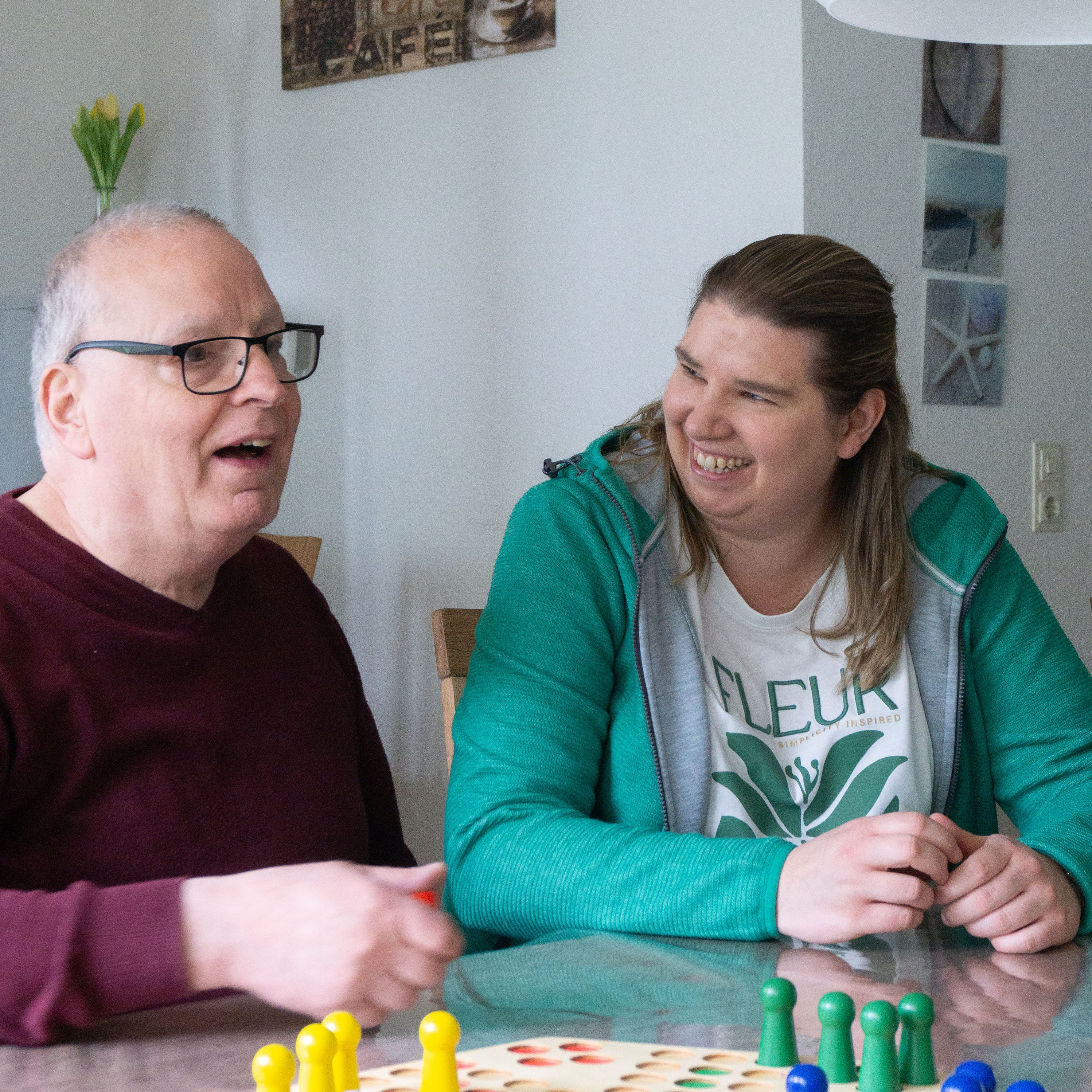Eine Mitarbeiterin und eine älterer Mann sitzen an einem Tisch und lachen. Vor ihnen ist ein "Mensch ärgere dich nicht"-Spiel auf dem Tisch zu sehen.