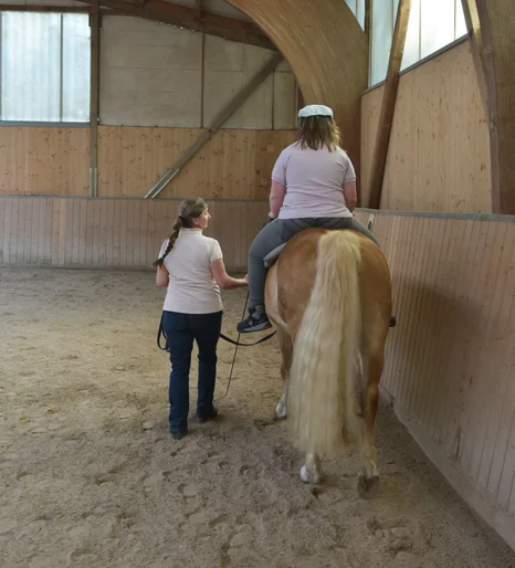 Eine Frau führt ein Pferd, auf dem ein Mädchen sitzt. Man sieht beide nur von hinten in einer Reithalle.