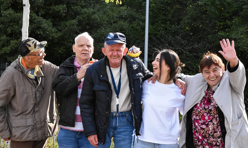 Eine Gruppe von Senioren und eine junge Frau halten sich in den Armen. Drei von ihnen lachen, eine Frau winkt. 