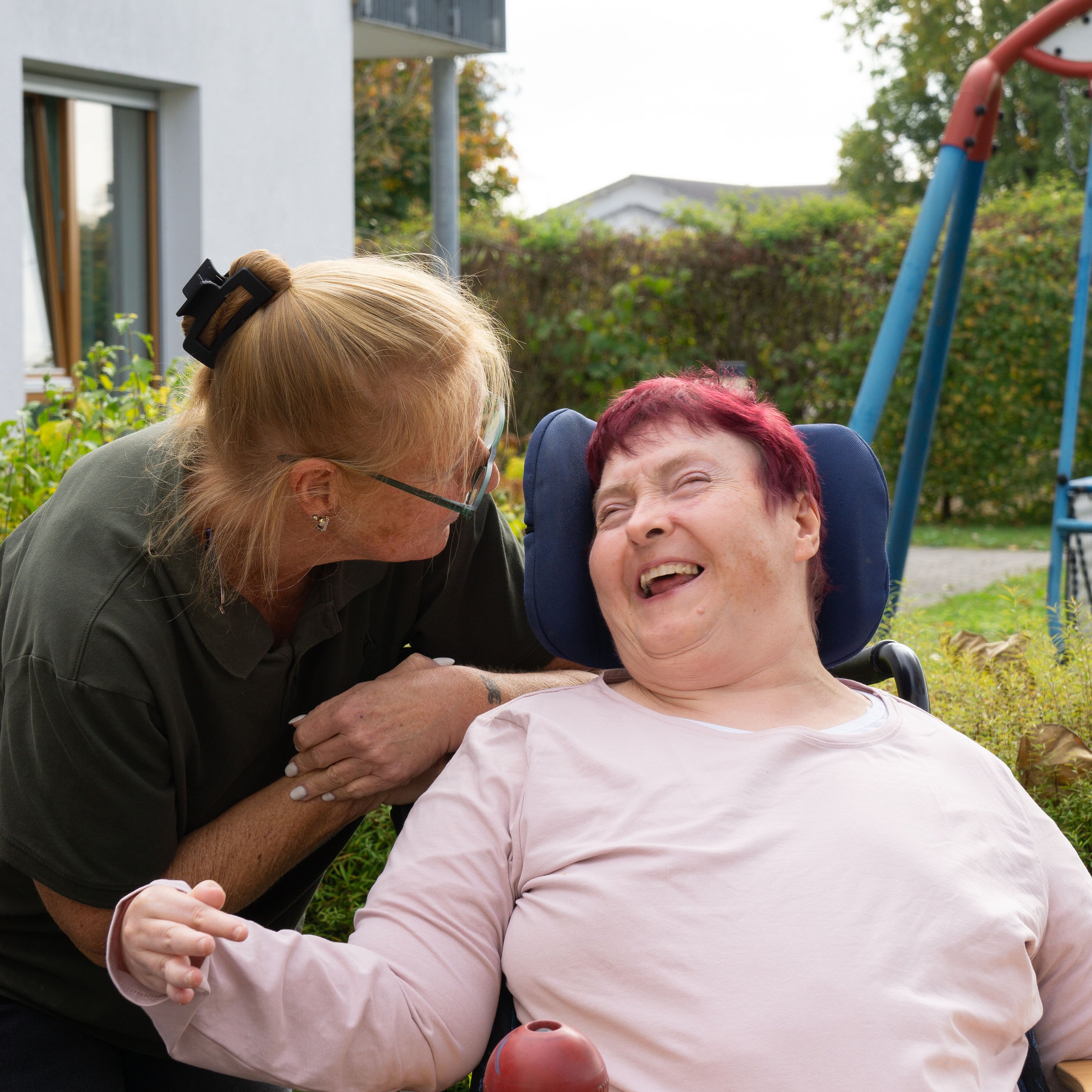 Eine Frau mit Behinderung sitzt in einem Rollstuhl draußen in einem Garten und lacht. Eine Mitarbeiterin beugt sich zu ihr und schaut sie an. 