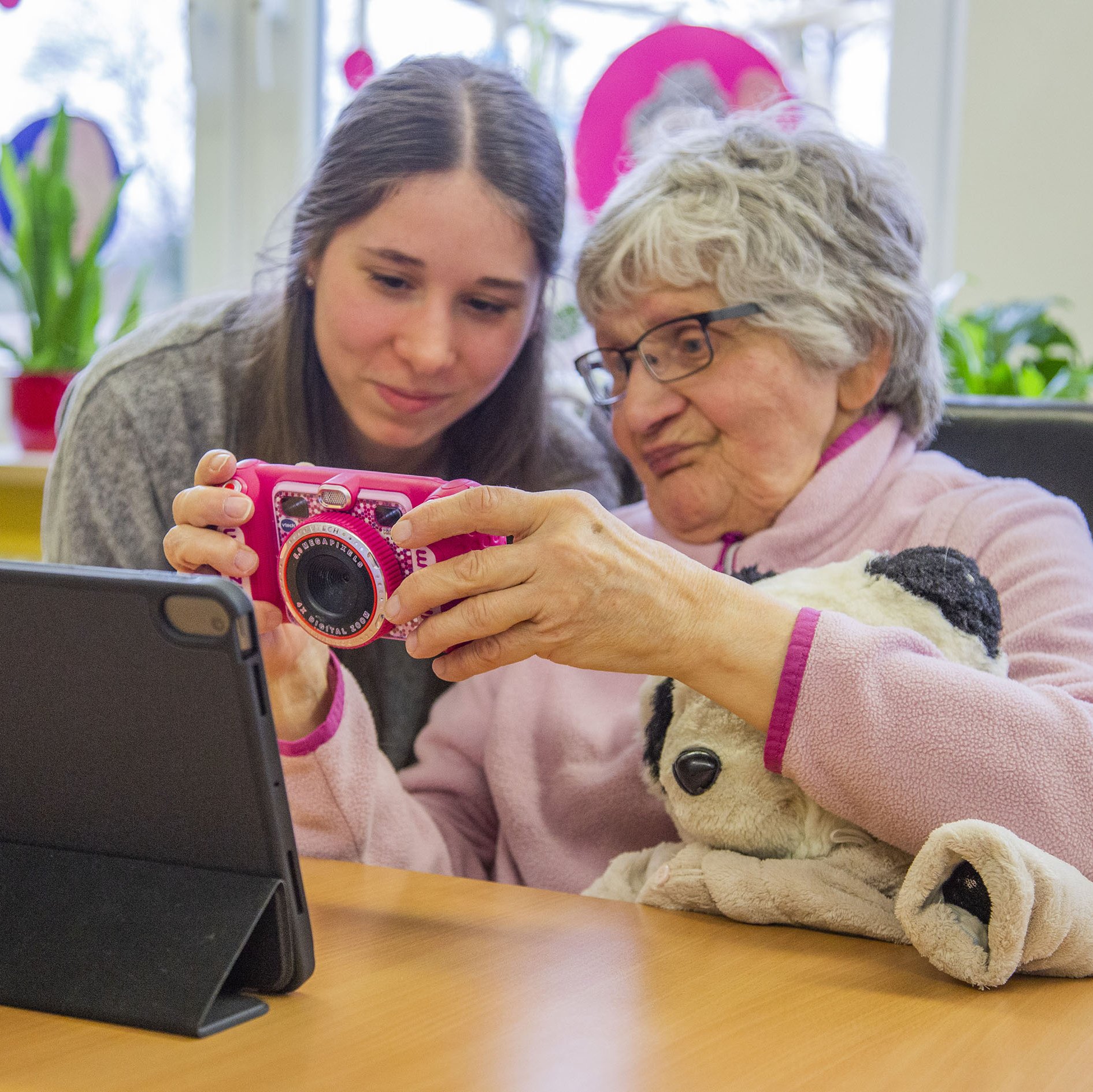 Eine ältere Frau zeigt einer jungen Frau ihre Kamera, sie sitzen an einem Tisch und vor ihnen steht ein Tablet auf dem Tisch.