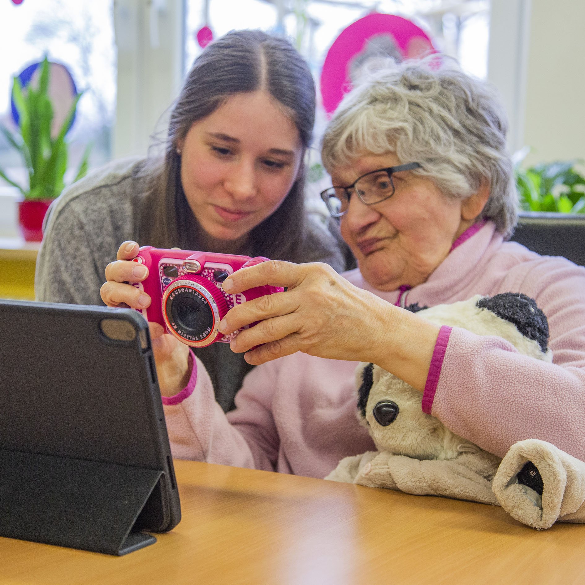 Eine ältere Frau zeigt einer jungen Frau ihre Kamera, sie sitzen an einem Tisch und vor ihnen steht ein Tablet auf dem Tisch.