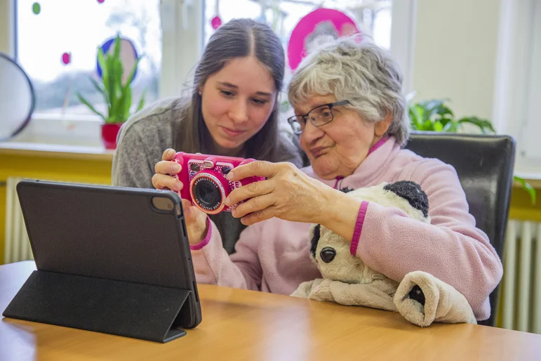 Eine ältere Frau zeigt einer jungen Frau ihre Kamera, sie sitzen an einem Tisch und vor ihnen steht ein Tablet auf dem Tisch.