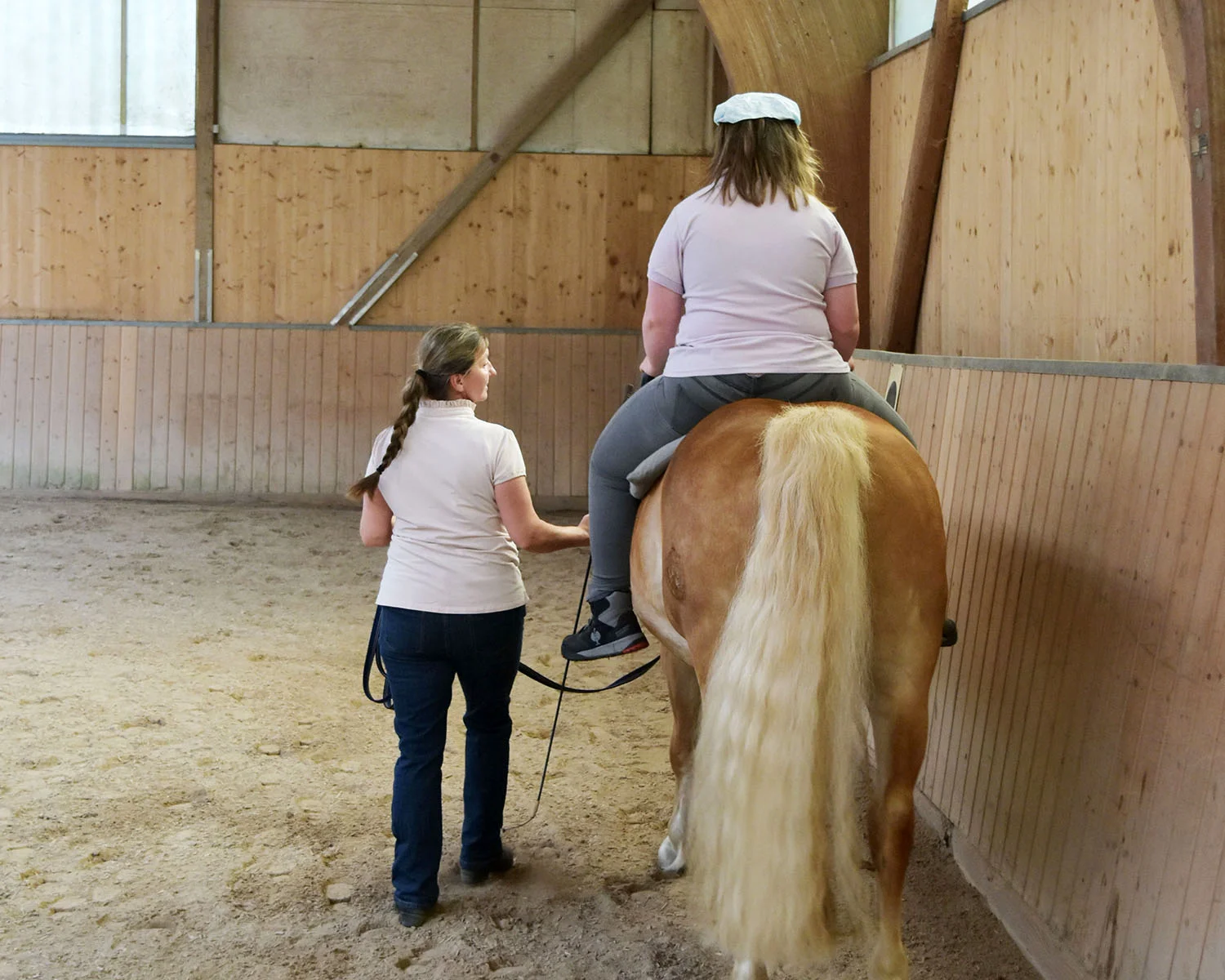 Eine Frau führt ein Pferd, auf dem ein Mädchen sitzt. Man sieht beide nur von hinten in einer Reithalle.