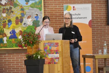 Zwei Frauen stehen hinter einem Rednerpult, an dem ein buntes Plakat der Fachtagung hängt. Eine Frau spricht.