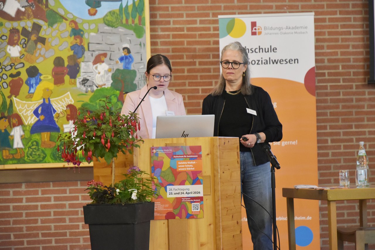 Zwei Frauen stehen hinter einem Rednerpult, an dem ein buntes Plakat der Fachtagung hängt. Eine Frau spricht.