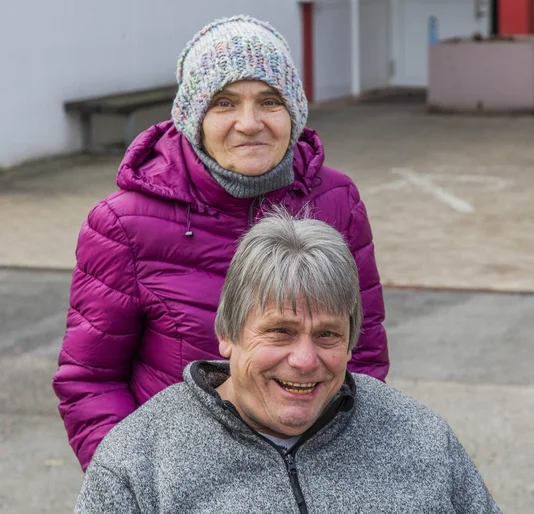 Eine Frau mit Jacke und Mütze steht hinter einem Mann mit grauen Haaren, dieser sitzt.