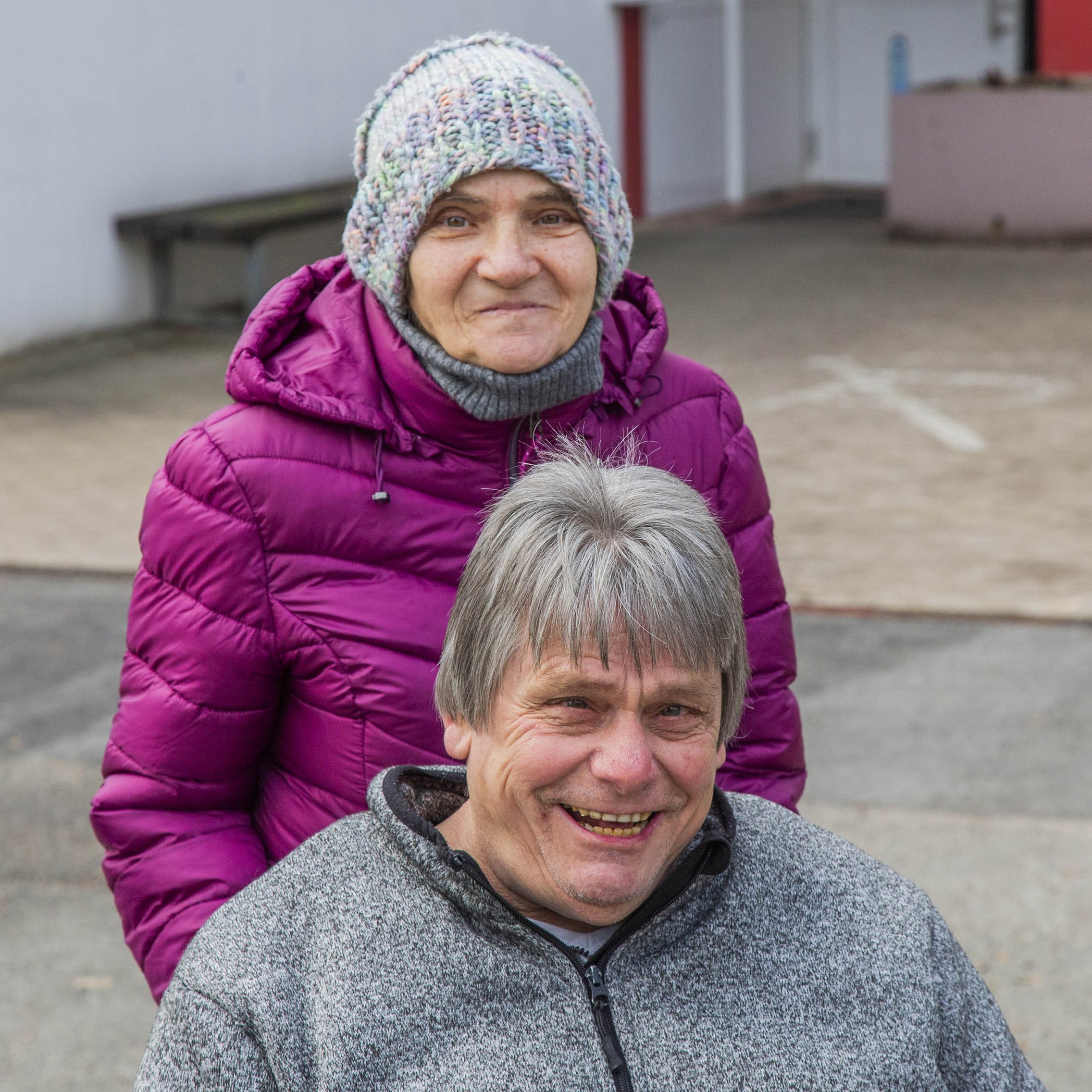 Eine Frau mit Jacke und Mütze steht hinter einem Mann mit grauen Haaren, dieser sitzt.