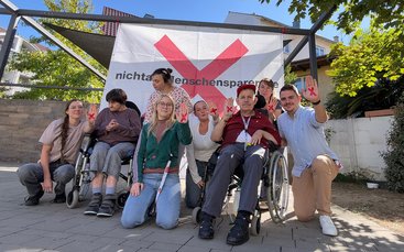 Bewohner, Bewohnerinnen und Mitarbeitende sitzen und knien vor einem Protestbanner.