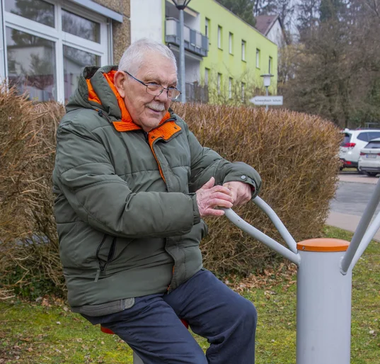 Ein älterer Mann mit Winterjacke sitzt auf einem Sportgerät draußen in der Natur.