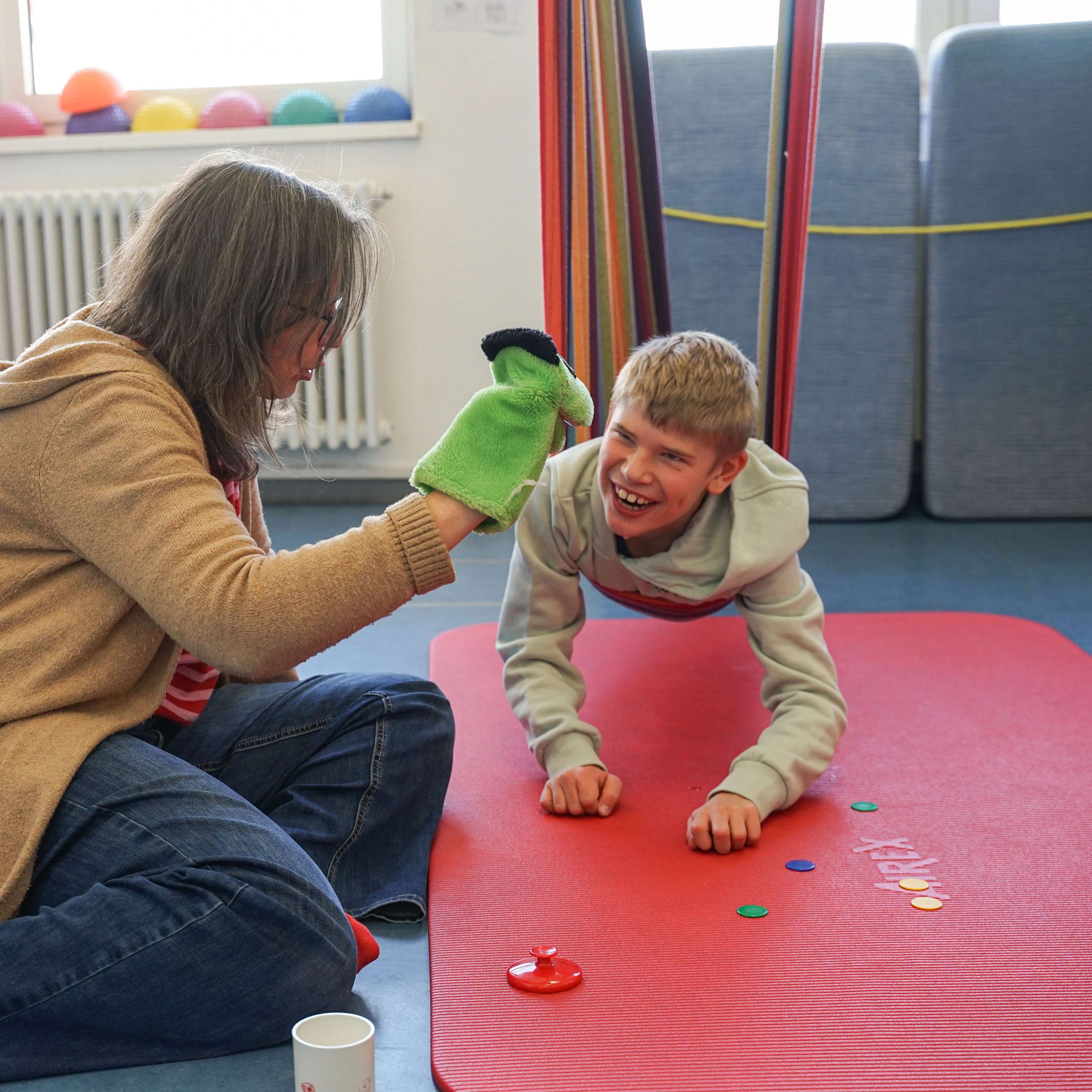 Ein Junge liegt auf dem Bauch in einer Tuch-Schaukel, die über einer Matte hängt. Eine Frau sitzt neben ihm und spielt mit einer Handpuppe.