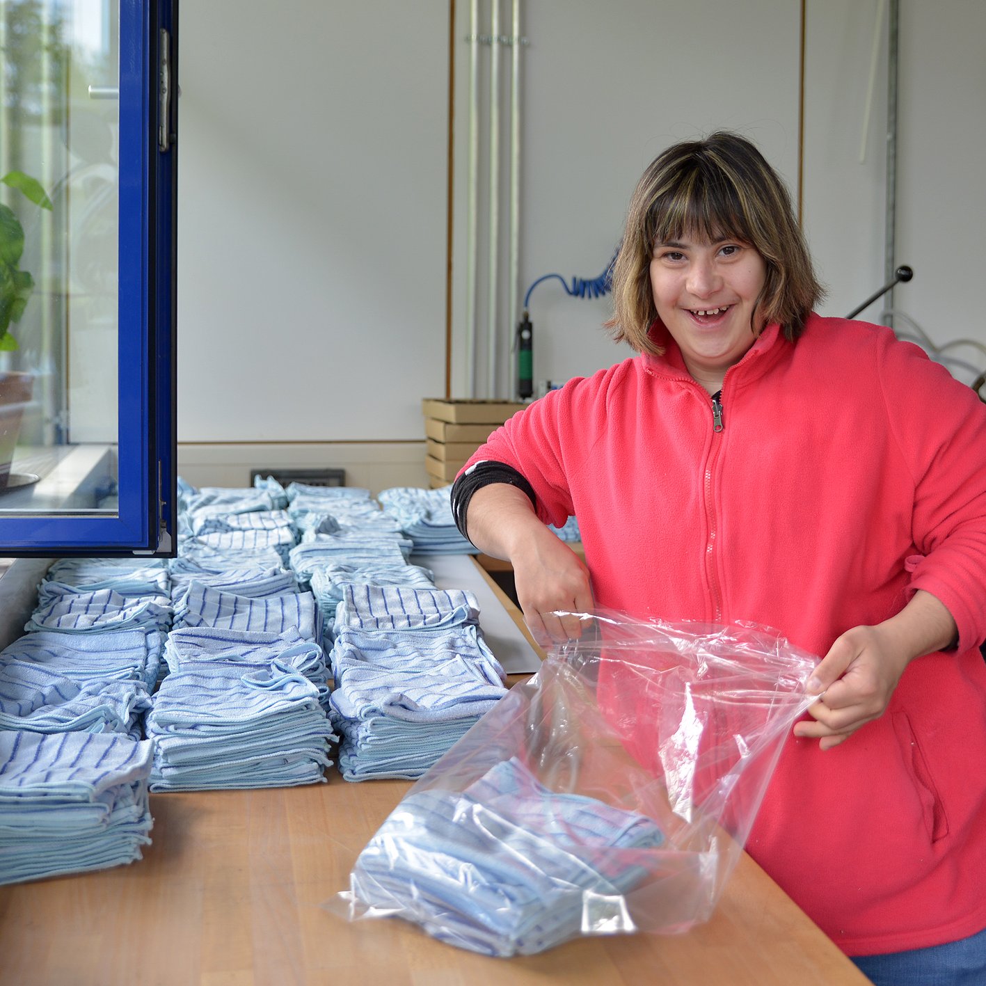 Eine Frau steht an einem Tisch, vor ihr liegen gefaltete Stoffe. Sie packt diese in eine Plastiktüte.