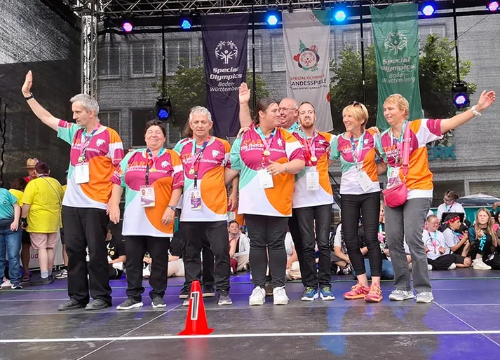 Acht Menschen in bunten Johannes-Diakonie Triktos jubeln auf einer Bühne der Special Olympics Landesspiele.