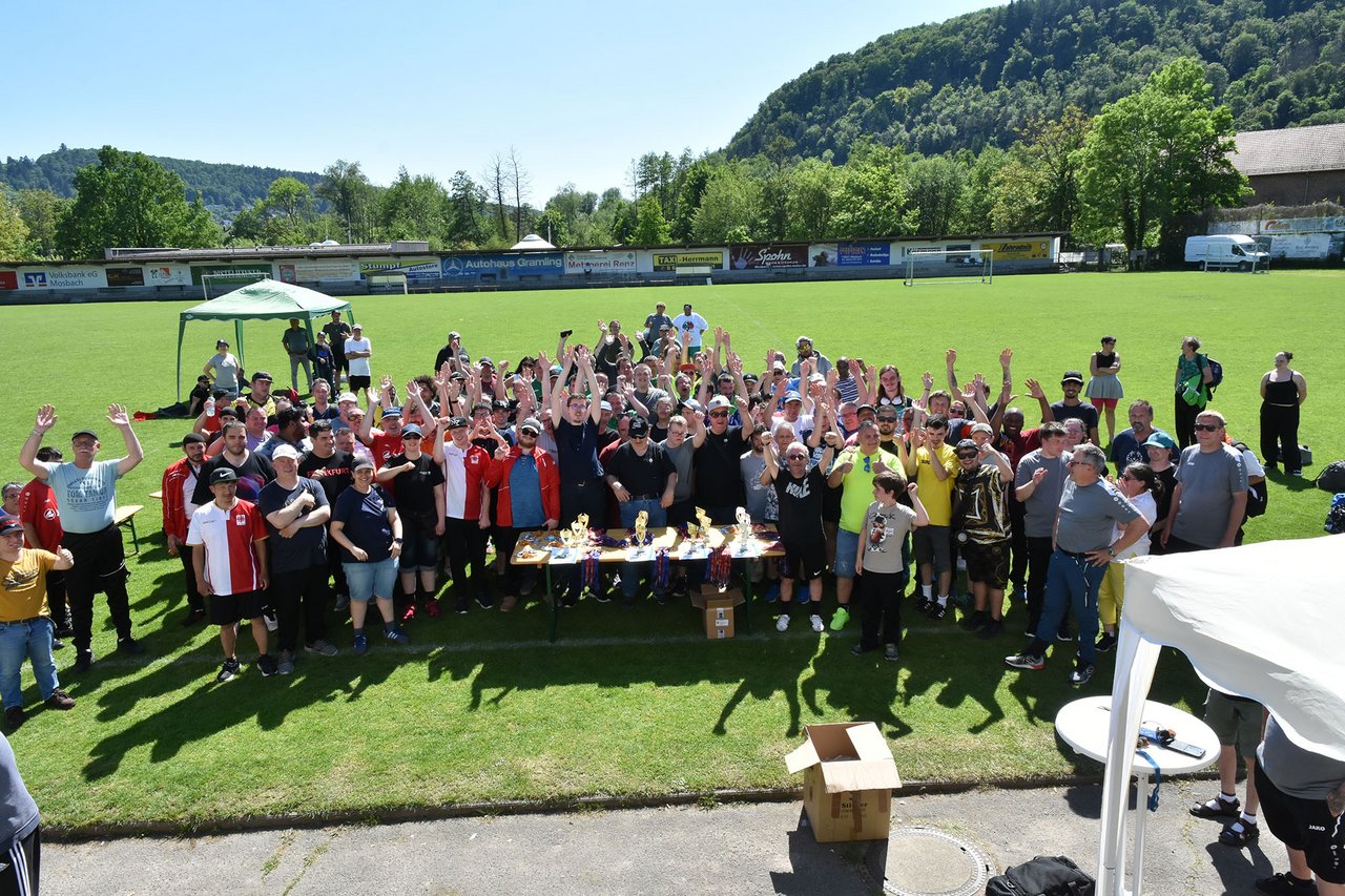 Gruppenfoto von allen Teilnehmenden Fußballteams und Trainer*innen auf dem Sportplatz. Die Menschen heben jubelnd ihre Arme. 