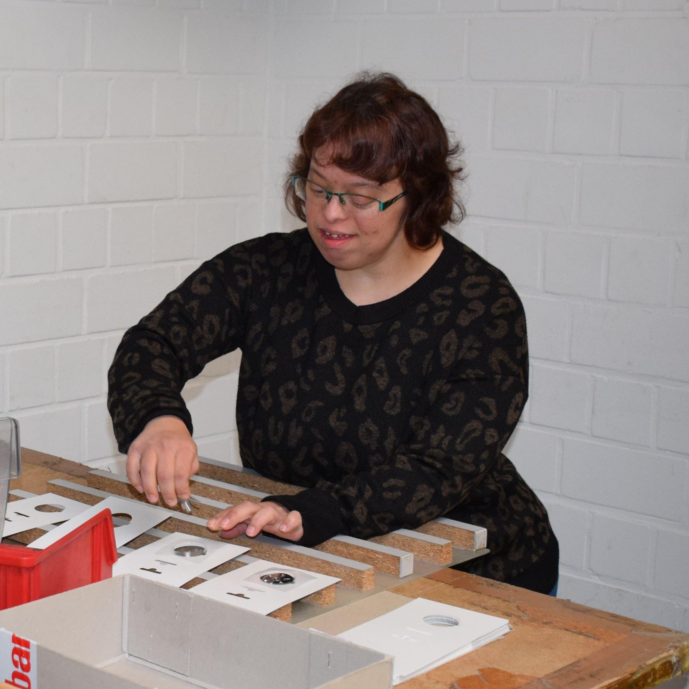 Eine Frau steht hinter einem Arbeitstisch mit verschiedenen Materialien und setzt diese zusammen.