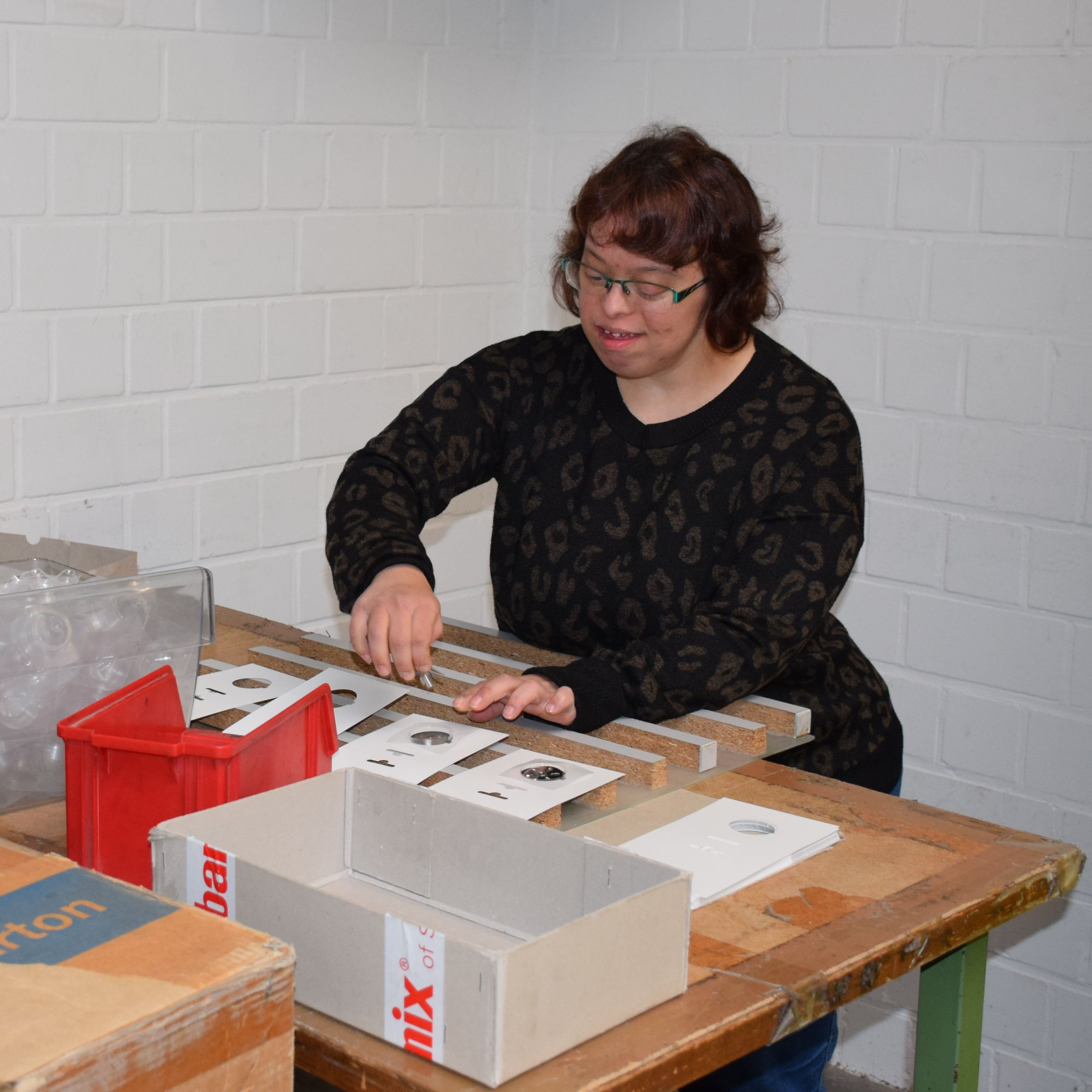 Eine Frau steht hinter einem Arbeitstisch mit verschiedenen Materialien und setzt diese zusammen.