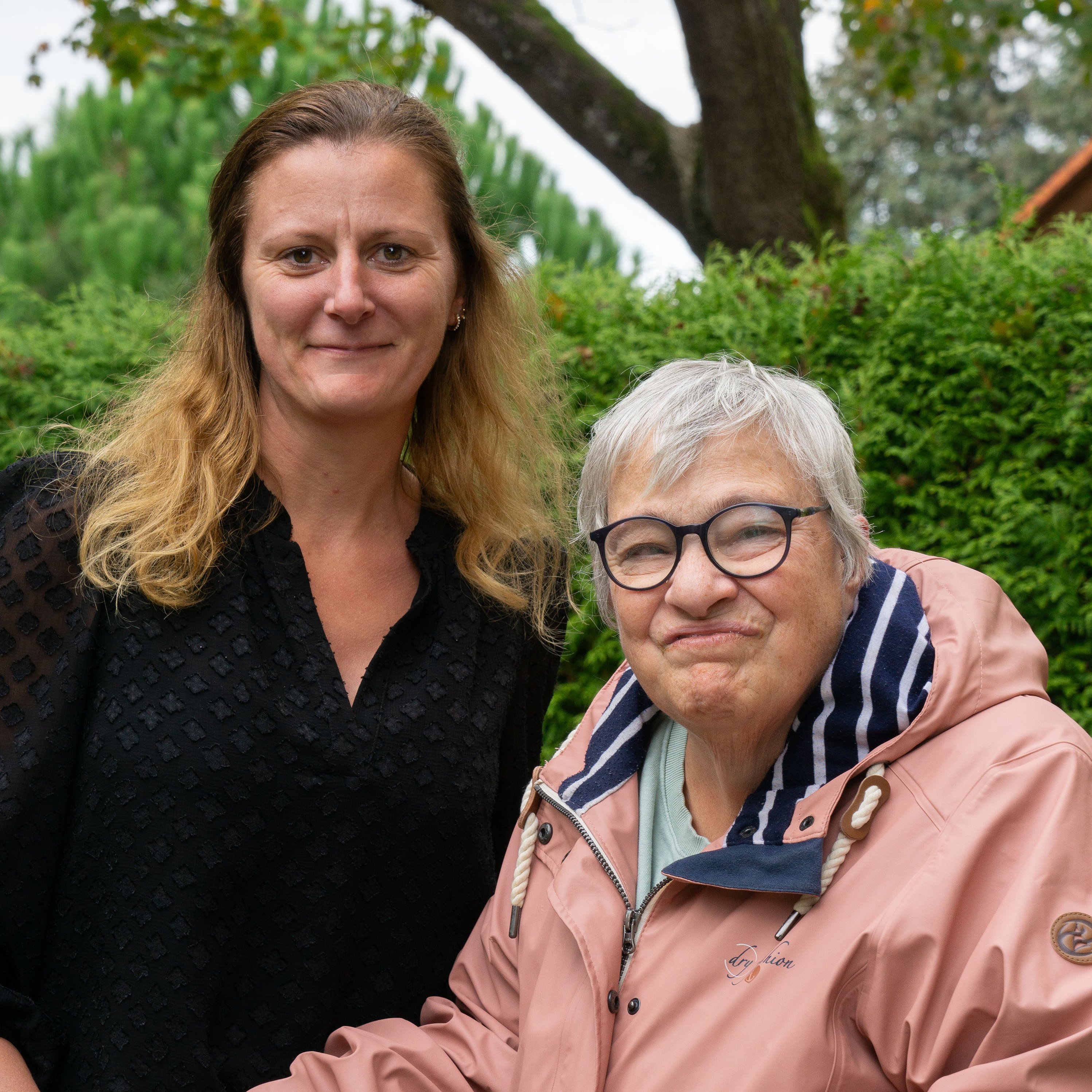 Eine Mitarbeiterin und eine ältere Frau mit Rollator stehen im Grünen und schauen in die Kamera.