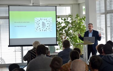 Volkhard Menzel steht vor Publikum neben einer Leinwand und hält einen Vortrag mit Infofolien.