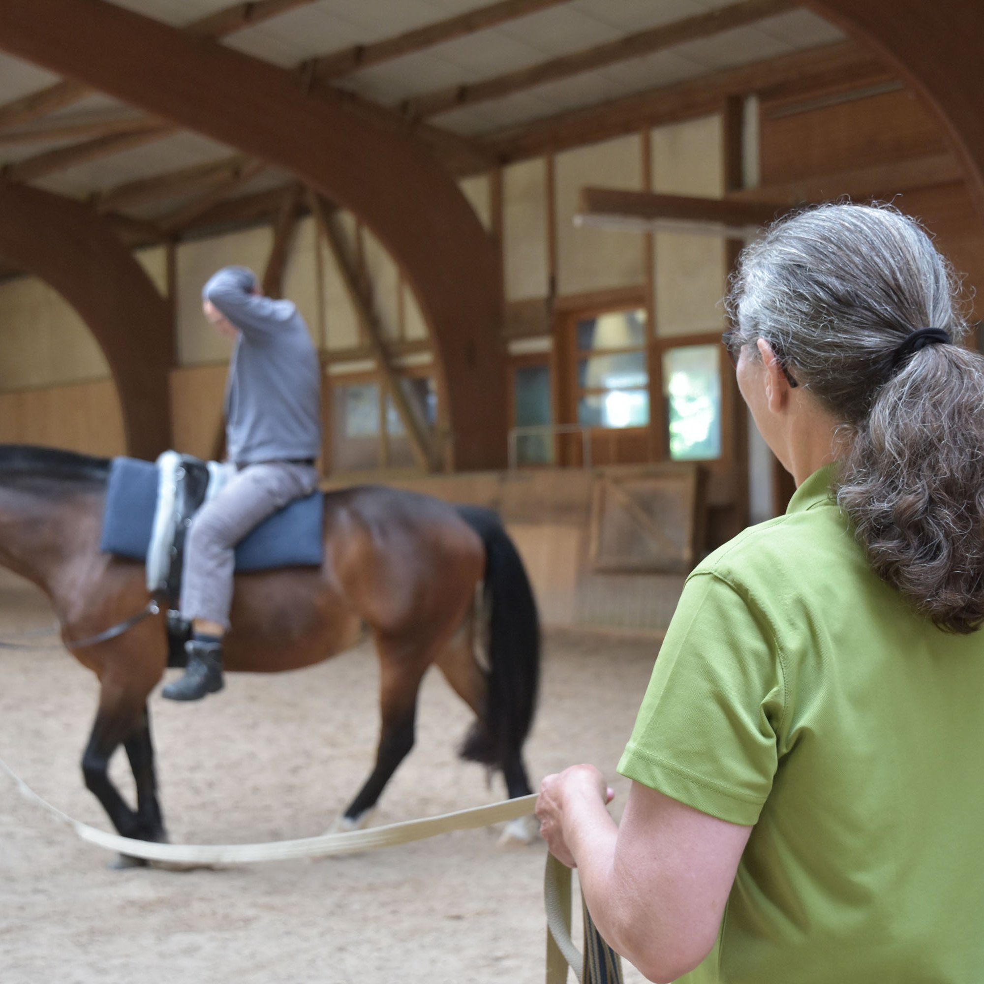In einer Reithalle reitet ein Mann auf einem Pferd, dieses wird von einer Frau an einer langen Leine geführt.