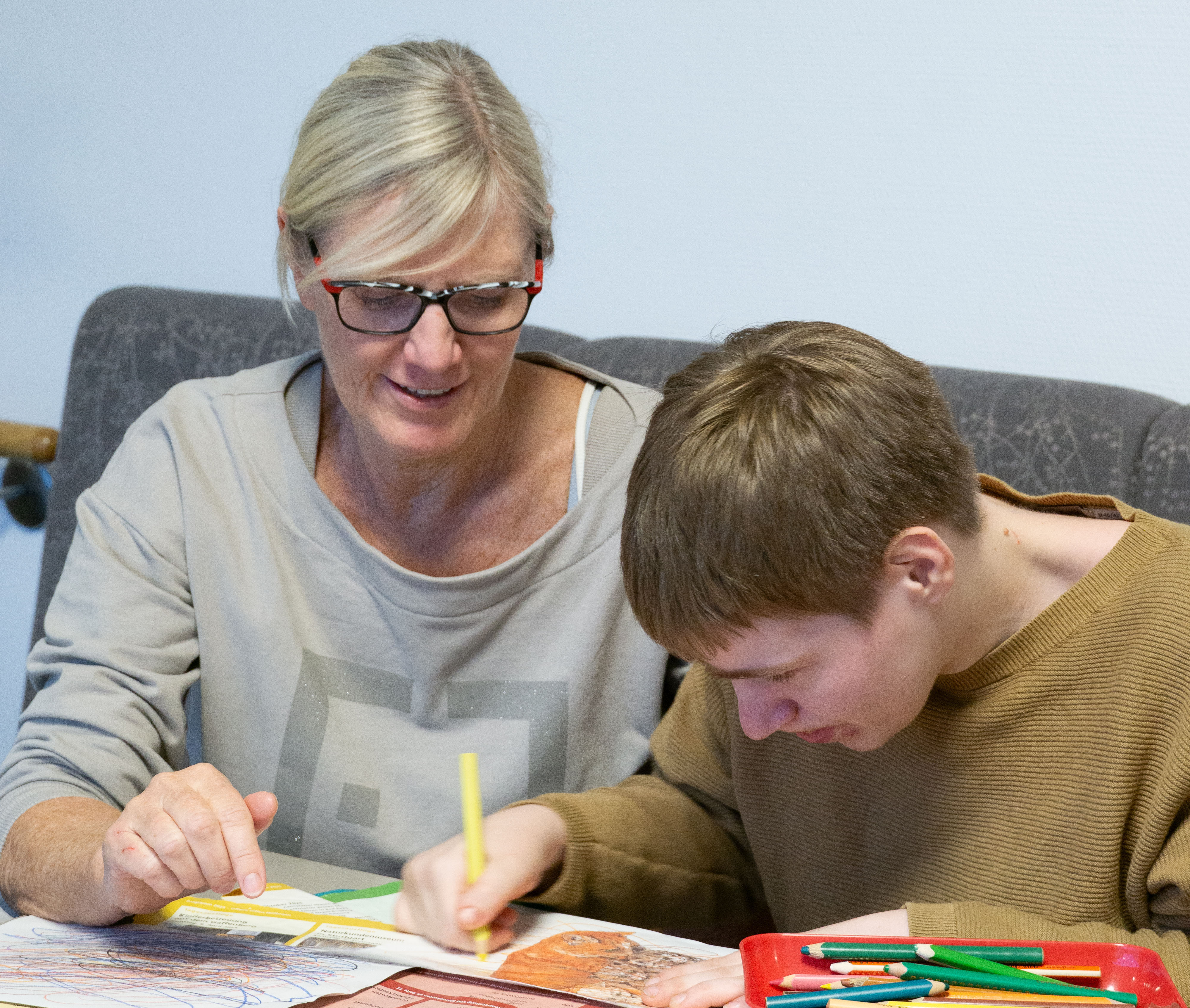Eine Frau unterstützt einen jungen Mann beim Malen, sie sitzen an einem Tisch.