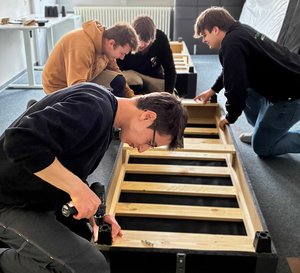 Auszubildende der Firma Mosca bauen mit Auszubildenden des Berufsbildungswerks Mosbach-Heidelberg in einem Zimmer Möbel zusammen.
