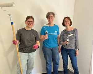 Drei SAP-Mitarbeiterinnen stehen mit Pinseln und Farbrollen vor einer frisch gestrichenen Wand.