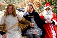Ein als Christkind junger Mann und ein Mann im Nikolauskostüm sitzen gemeinsam mit einer Frau auf einer Bank im Freien und lachen fröhlich in die Kamera.