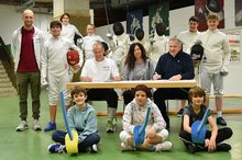 Zwei Männer und eine Frau sitzen an einem Tisch, umgeben von Kindern und Jugendlichen mit Fechtausrüstung. 