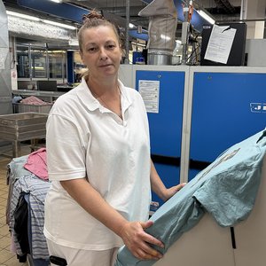 Eine Frau steht in einer Großwäscherei an einem Gerät zum Glätten von Hemden und schaut in die Kamera.